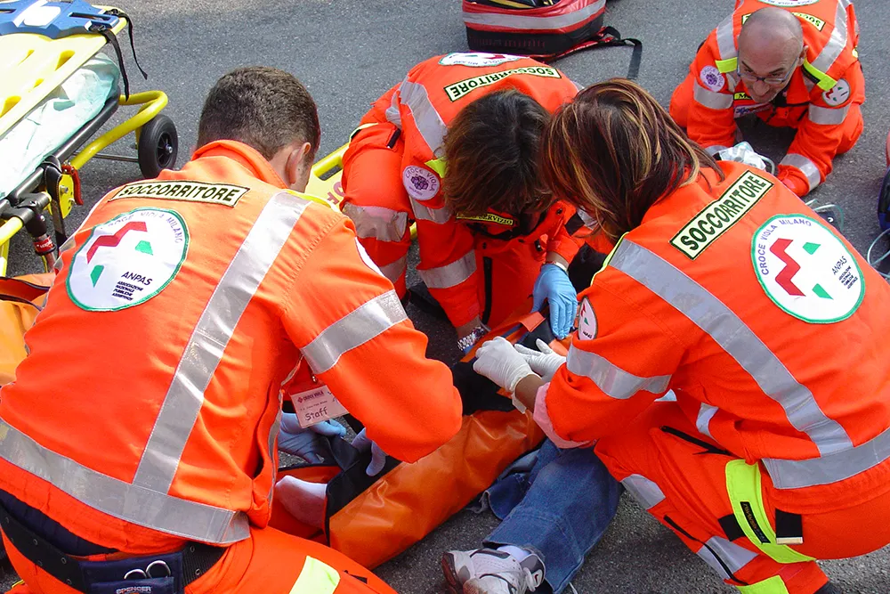 Squadra di soccorritori in divise arancioni ad alta visibilità presta primo soccorso a persona infortunata su asfalto, con barella e attrezzatura medica.