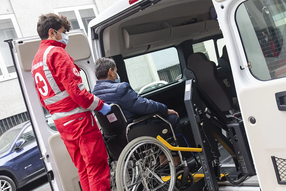 Volontario della Croce Rossa in divisa rossa assiste paziente in sedia a rotelle salendo su ambulanza bianca con pedana di accesso.