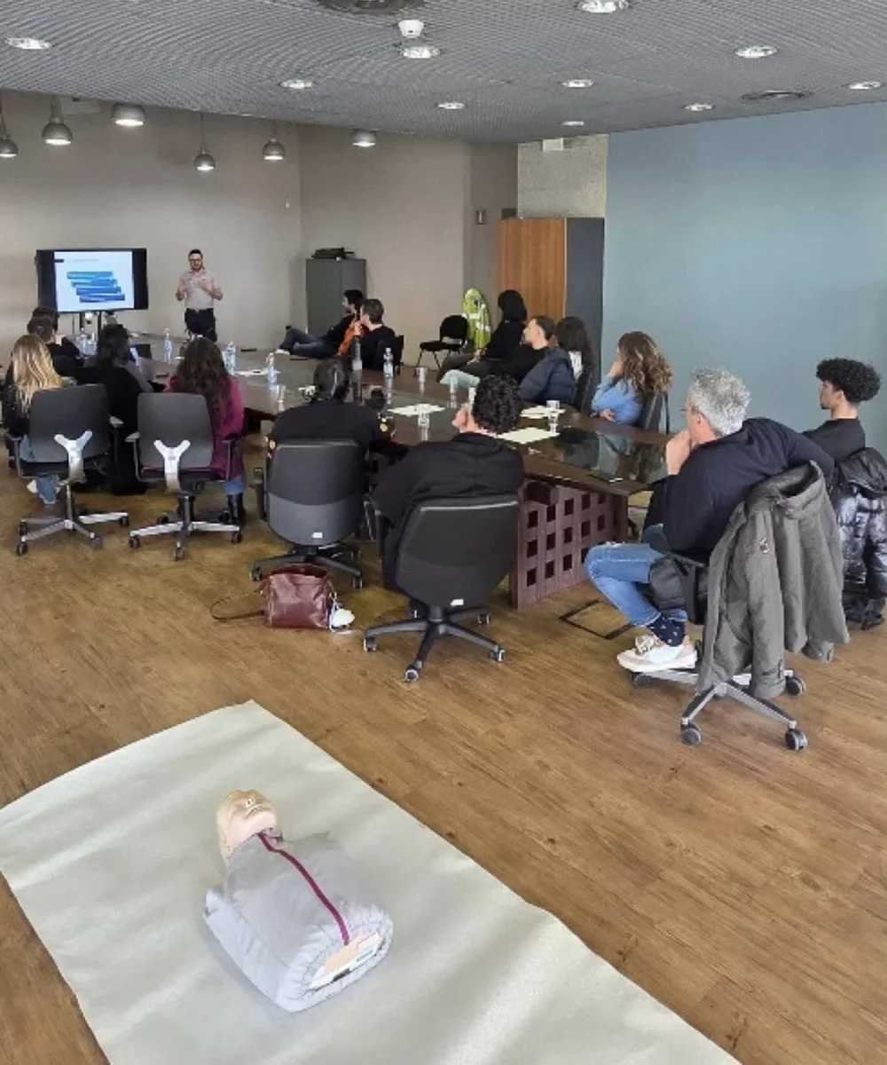 Corso di primo soccorso in sala conferenze: formatore davanti a uno schermo con presentazione, partecipanti seduti ai tavoli e manichino per la rianimazione in primo piano