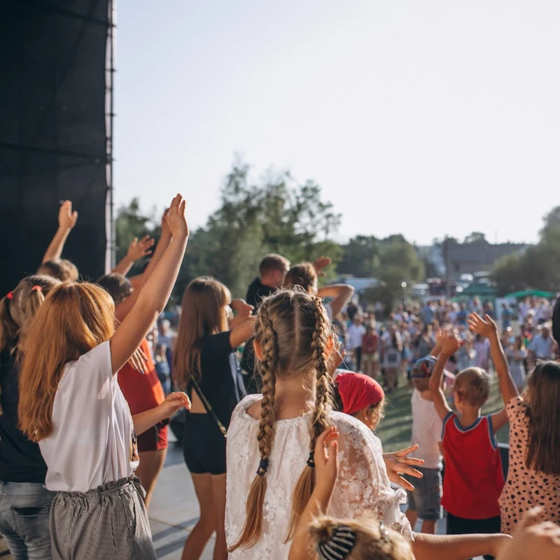 Giovani partecipanti a festa o concerto estivo alzano le mani con energia davanti a palco, atmosfera gioiosa in spazio verde pubblico.