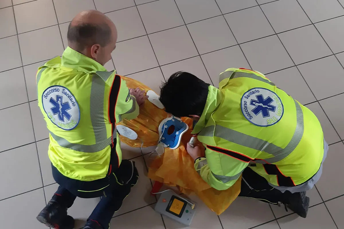 Operatori sanitari con divise riflettenti gialle e bande grigie applicano dispositivi di immobilizzazione su paziente disteso su asse rigida color arancio, defibrillatore giallo visibile accanto, scena di soccorso professionale vista zenitalmente.