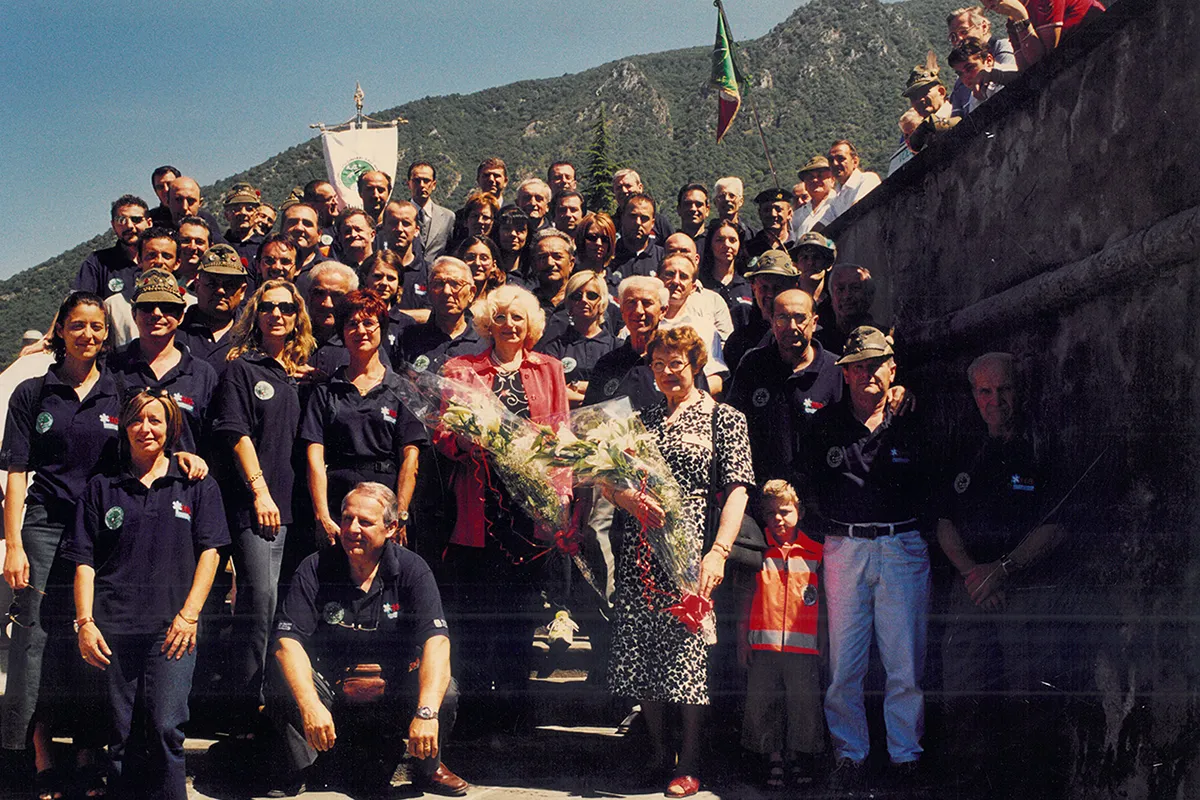 Foto d'archivio di raduno associativo con volontari soccorritori in divise operative blu navy, alcuni con cappelli da Alpini, e rappresentanti femminili in abiti da cerimonia con mazzi floreali, vessillo verde associativo visibile, scalinata monumentale e paesaggio prealpino come scenario naturale.