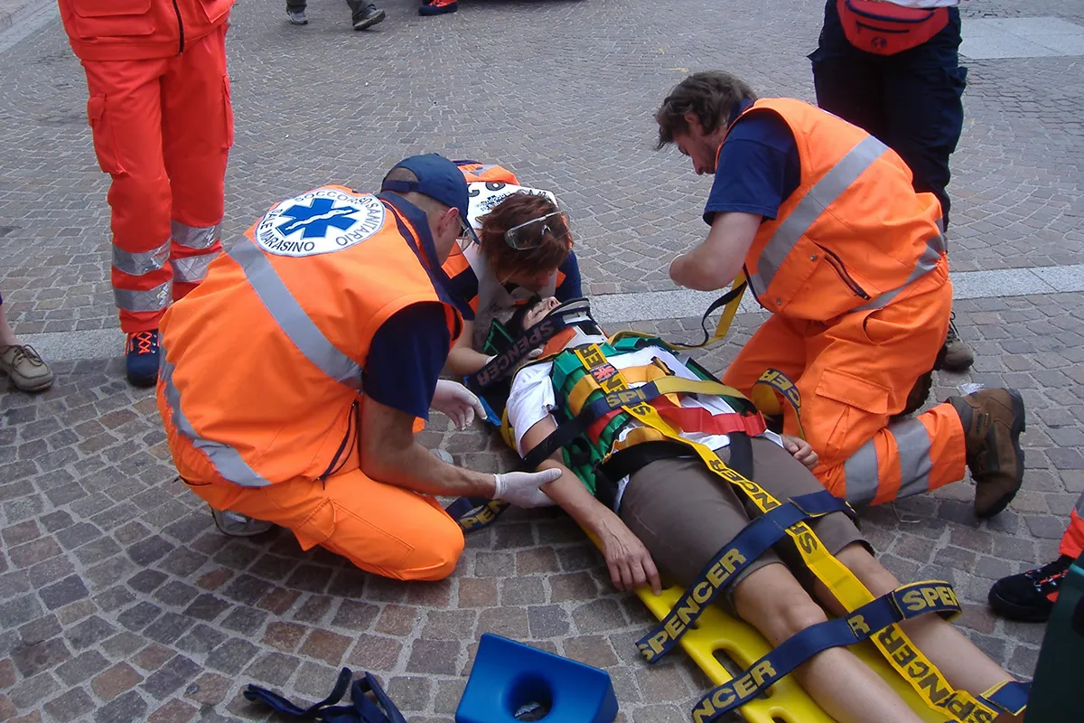 Team di volontari protezione civile in uniformi arancio fluorescente con bande riflettenti applica dispositivi di stabilizzazione spinale su persona con collare cervicale e blocchi laterali per testa, simulazione intervento emergenza in piazza.