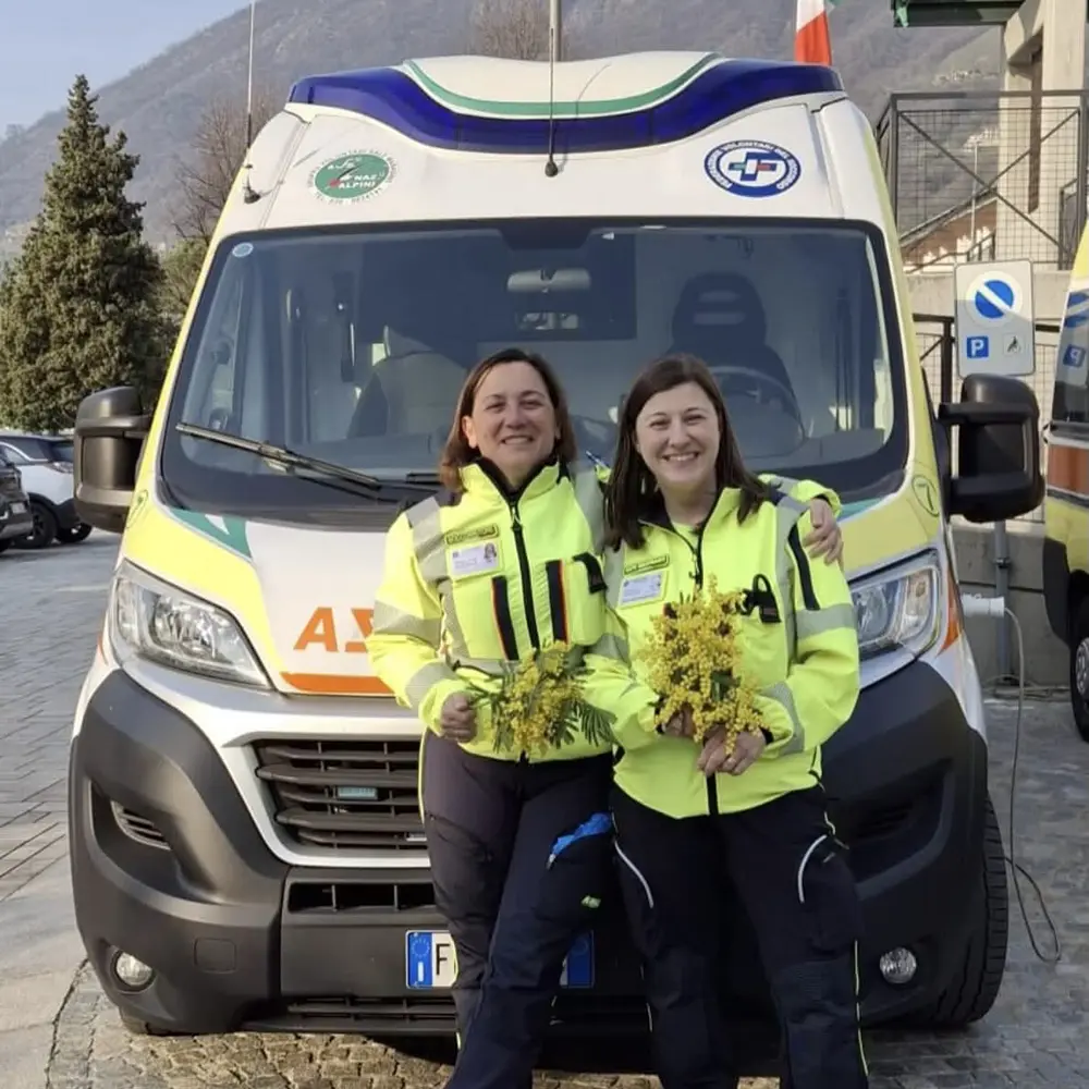 Due volontarie in divisa gialla fluorescente sorridenti davanti all'ambulanza, con in mano mazzi di mimosa per la Festa della Donna