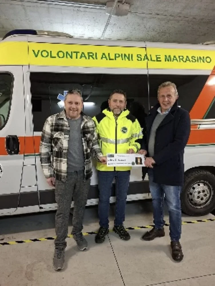 Un volontario in divisa gialla riceve un assegno di donazione da due uomini davanti all'ambulanza dei Volontari Alpini Sale Marasino