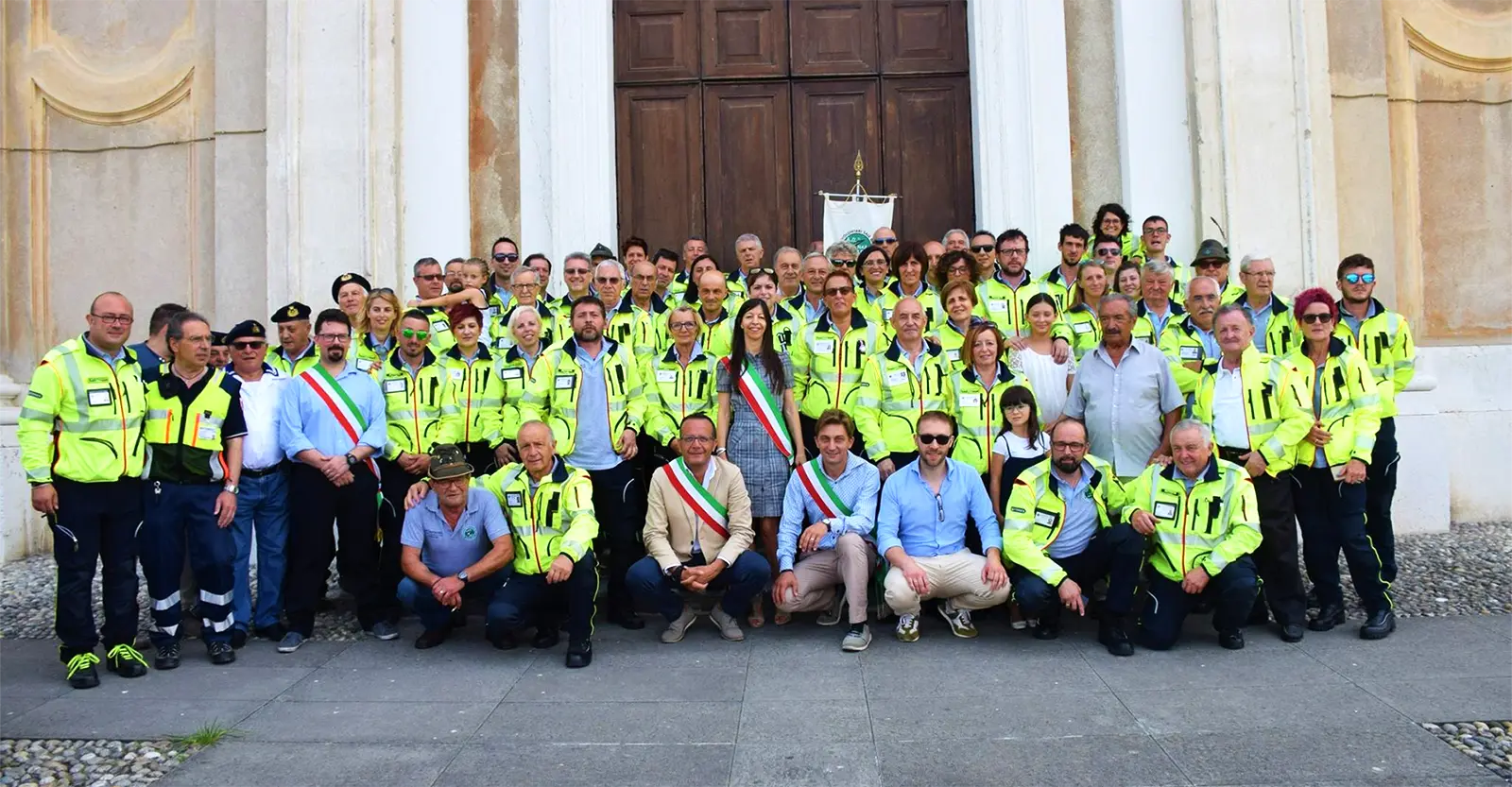 Numerosi volontari del soccorso in giacca alta visibilità riuniti in foto ufficiale davanti a una chiesa, insieme al sindaco e alle autorità locali con fascia tricolore.