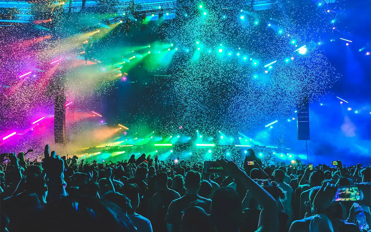 Folla numerosa di persone a un concerto con le mani alzate davanti a un palco illuminato da luci colorate verde blu e rosa. Coriandoli e fumo riempiono laria mentre il pubblico riprende lo spettacolo con i propri smartphone.